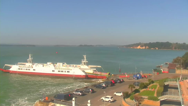 The image shows a ferry docked at a harbor on a sunny day. The ferry, primarily white with red accents, is positioned near a concrete pier. Surrounding the ferry, there are several parked cars and some palm trees in the foreground. In the background, calm blue water extends toward a distant shoreline with trees and cliffs. A clear blue sky is visible above.