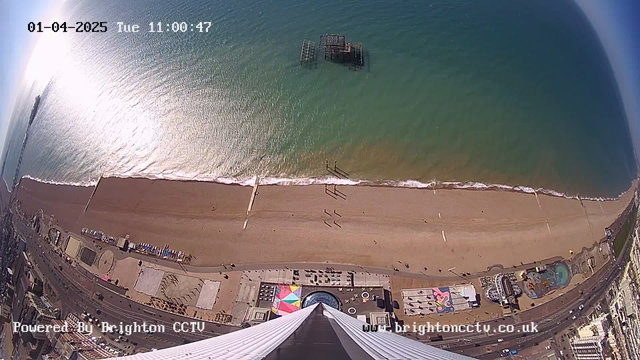 An aerial view of a beach with golden sand and gentle waves lapping at the shore. The ocean appears turquoise, reflecting sunlight. People can be seen walking along the beach. In the distance, an old pier emerges from the water. Various beachside attractions, including colorful structures and pathways, are visible on the sand. The scene is captured from a high vantage point, showcasing the coastline and surrounding area. The image is timestamped and labeled as powered by Brighton CCTV.