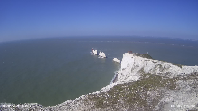 A clear blue sky stretches above the ocean, with gentle waves visible. In the foreground, white chalk cliffs rise steeply from the water, while small red and white lighthouse structures sit on rock formations in the sea. The coastline appears rugged and natural, dotted with patches of grass and earth. The scene conveys a sense of openness and tranquility, with no visible land beyond the cliffs and lighthouses.