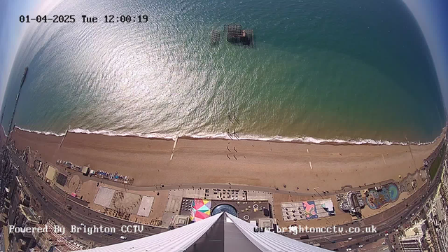 Aerial view of a beach scene on a sunny day. The ocean is calm with waves gently lapping at the shore. Below, a wide expanse of sandy beach is visible, dotted with small figures of people walking along the water's edge. To the left, there is a pier extending into the ocean, with a structure visible on the water. Various beach amenities and colorful installations can be seen, including umbrellas and a large circular structure on the beach. The image captures a bright blue sky overhead, adding to the vibrant atmosphere.