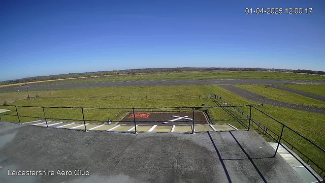 A clear blue sky with no clouds is visible above an airport landscape. In the foreground, there is a rooftop railing with a flat surface below, leading to a helipad marked with a large white cross on a red background. Beyond the helipad, a grassy area and a runway are visible, bordered by wooden fencing. The scene is well-lit, indicating it is daytime.