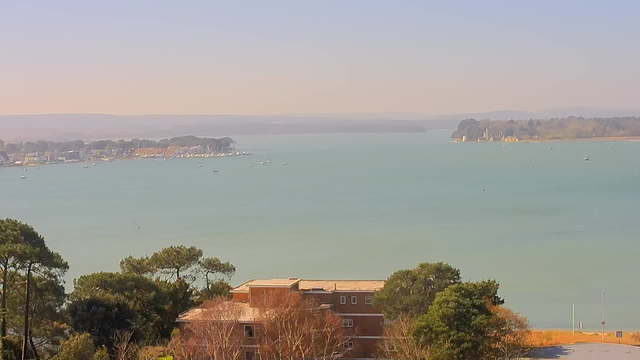A scenic view of a calm bay under a clear sky. In the foreground, there are some trees and a building with a flat roof. The water appears still and is a light blue-green color, with several small boats scattered across the bay. In the distance, a shoreline lined with buildings extends along the water's edge, leading to a wooded area and a small island with what looks like a historical structure. The background features gentle hills fading into the horizon.