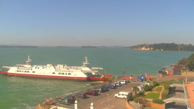 A ferry boat is docked at a marina with vehicles parked nearby. The scene shows a calm body of water under a clear blue sky, with some distant land visible on the horizon. A few people can be seen near the ferry, and there are clusters of parked cars in various colors along the waterfront. The landscape includes manicured green areas and pathways leading towards the water.