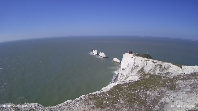 A clear blue sky stretches overhead, melding into the ocean below, which is a greenish blue. In the foreground, there is a rocky cliff with patches of grass and stones. Prominent white chalk cliffs rise from the shoreline, with two distinct white rock formations jutting out into the sea. A red lighthouse stands atop one of the formations, and a small building is visible on top of the right cliff. The overall scene conveys a bright and sunny coastal landscape.