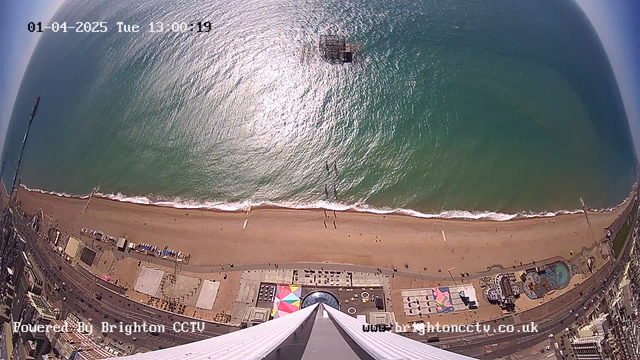 Aerial view of a beach. The image shows a sandy shoreline meeting the ocean, with gentle waves lapping at the sand. In the foreground, there is a variety of beach facilities, including colorful structures, likely cafes or attractions. A distant pier extends into the ocean, partially visible, and scattered people can be seen walking along the beach and boardwalk. The scene is sunny with clear skies, reflecting bright light on the water's surface. At the bottom of the image, there is a partially visible structure, possibly a building, with large angular lines leading to the camera's viewpoint. The date and time are displayed in the top corner.