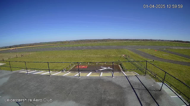 A clear blue sky stretches overhead with no clouds. In the foreground, there is a flat rooftop with black railings. Below, a grassy area features a large white "X" marking on the ground, surrounded by a rectangular patch of reddish material. The background shows a runway with marked lines, and further back, there are fields and low rolling hills. The scene indicates a daytime view from the Leicestershire Aero Club.