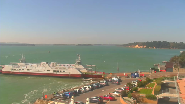 A view of a harbor with a large ferry docked at the pier, surrounded by calm turquoise waters. The sky above is clear and blue, with a few distant islands visible on the horizon. Along the shoreline, a parking area filled with various cars is seen, with some greenery and walkways leading towards the water. The scene depicts a peaceful day at the waterfront.