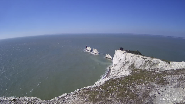 A panoramic view of a coastline featuring white chalk cliffs against a clear blue sky. In the distance, several rock formations emerge from the ocean, with a small red lighthouse visible on one of the rocks. The water appears calm with gentle waves, reflecting the sunlight. The foreground shows green grass and rocky terrain leading to the cliff edge.