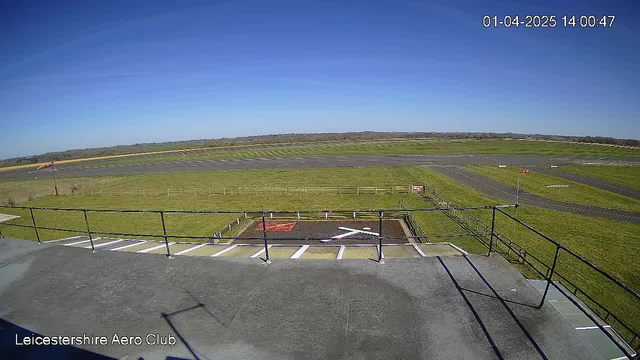 A clear blue sky stretches across the top of the image. Below, there is a grassy area adjacent to an airstrip, which is marked by parallel lines. In the foreground, a flat rooftop features a safety railing and has a large 'X' marking on dark soil, indicating a landing or takeoff area, with a red area nearby. A striped windsock is visible to the left of the airstrip, indicating wind direction. The scene is bright and well-lit, suggesting daytime.