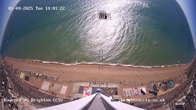 Aerial view of a sandy beach with a clear blue sea. The shoreline is lined with people enjoying the beach. In the distance, a pier extends into the water, partially submerged. Below, there are colorful structures and outlines of buildings, including a pool area and various attractions. The sky is mostly clear and sunny.