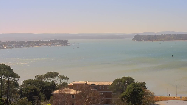 A serene waterfront view on a clear day, featuring calm turquoise water extending into the distance. The shoreline is lined with small boats and a few scattered buildings, partially obscured by trees in the foreground. Gentle hills are visible on the horizon, and the sky above is bright and mostly clear.