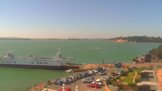 A scenic view of a harbor on a sunny day. In the foreground, there is a large ferry docked at the edge of a concrete pier. Several cars are parked in a lot near the pier, with a mix of colors and sizes visible. The water is a vibrant green, with gentle waves and small boats in the distance. In the background, there are tree-covered hills and sandy shores along the coastline, under a clear blue sky.