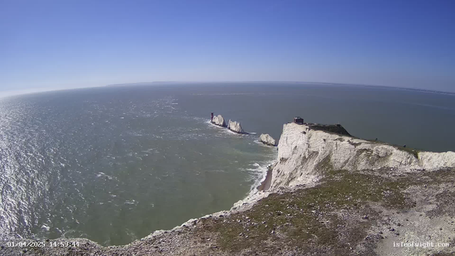 A wide view of a coastal scene featuring steep white chalk cliffs on the right, with a grassy area at the top of the cliffs. In the distance, several rock formations jut out into the sea. Below, the ocean stretches out in varying shades of blue and green, with gentle waves creating ripples on the surface. The sky above is clear with a few wispy clouds, indicating a sunny day.