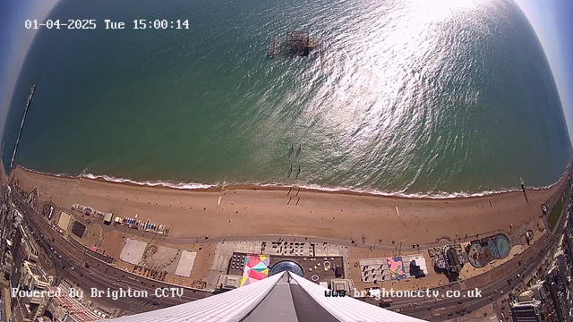 Aerial view of a sandy beach alongside the sea, taken from a high vantage point. The coastline is visible, with a few beachgoers walking along the shore. In the water, there is an old pier structure. The beach area features a colorful beach umbrella setup, several buildings, and attractions visible along the shore. The sky above is clear with the sun reflecting on the water surface, creating sparkles. The date and time are displayed in the upper left corner, indicating it's a Tuesday afternoon.