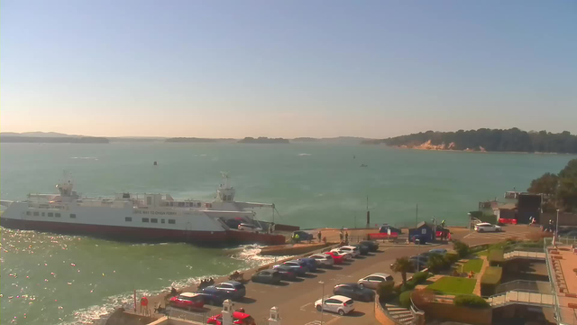 A ferry is docked at a pier in a waterfront scene with calm turquoise water. In the foreground, there is a parking lot filled with various cars. Some people are gathered near the ferry, and a few small boats can be seen on the water. The background features green hills and sandy cliffs under a clear blue sky.
