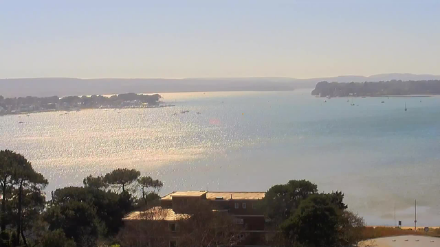 A serene coastal scene featuring a calm body of water reflecting sunlight, creating a shimmering effect. In the background, faint outlines of distant hills are visible. Several boats are scattered across the water, while trees and buildings line the shore in the foreground. The sky is clear, suggesting a bright day.
