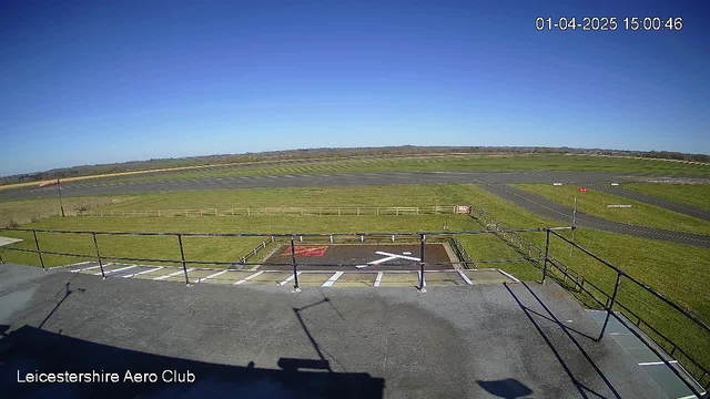 A clear blue sky stretches overhead, with no clouds visible. Below, a grassy area surrounds a tarmac airstrip, which has a mix of paved sections and grassy patches. A helipad is marked on the ground with a white 'H' symbol. Fencing borders the grassy areas, and there are some trees visible in the distance. The scene captures a tranquil moment at an aerodrome, showcasing open space and the airstrip's layout. The image is dated and time-stamped in the corner.