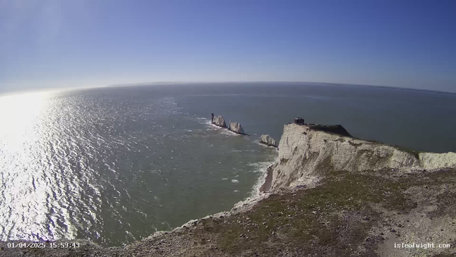 A scenic view of a coastline with white chalk cliffs. The cliffs rise sharply from the ocean, showcasing rocky formations jutting into the water. Below, there is a smooth beach area along the shore. The sky is clear and bright blue, while the water is shimmering with gentle waves reflecting the sunlight. In the distance, there are several rocky outcrops and a lighthouse structure. The overall atmosphere is tranquil and picturesque.