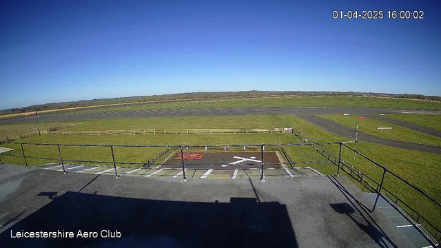 A clear blue sky is overhead, with a few vibrant green grassy fields visible below. In the foreground, there is a fenced area with an "X" marking on the ground, indicating a landing spot. An airstrip stretches out in the background, featuring a paved runway with markings. The scene conveys a wide, open landscape typical of an aviation area. In the distance, there are some trees lining the horizon. The time displayed is 16:00 on April 1, 2025.