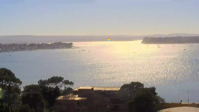 A serene coastal view featuring a vast body of water reflecting sunlight, creating a shimmering effect. In the distance, several small boats are scattered across the water, with a coastline visible on the left, lined with trees and buildings. The sky is blue with a slight gradient towards the horizon, suggesting a peaceful, sunny day.