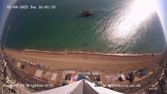 A wide aerial view of a beach and ocean scene captured from a high vantage point. The sandy beach stretches along the lower part of the image, with small figures walking near the shoreline. In the water, gentle waves are visible. A pier, partially submerged in the water, is situated toward the center. The beach is lined with various geometrical structures and colorful patterns visible on the ground. Above, the sky is mostly clear with a bright sun reflecting on the ocean surface, creating a shimmer effect. The timestamp on the image indicates it is April 1, 2025, at 4:01 PM.