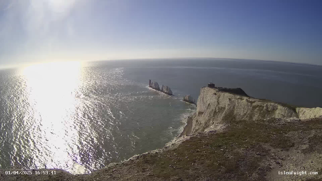 A wide view of the coastline featuring steep white cliffs on the right side, leading down to the sea with gentle waves. In the distance, several rock formations are prominent, with one rock having a tall, red structure atop it. The water reflects sunlight, creating a shimmering effect, while the sky is clear with a few wispy clouds. The image indicates a sunny day.