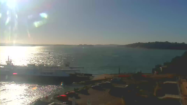 A scenic view of a harbor during the day. The sun shines brightly in a clear blue sky, casting light on the shimmering water. A large white boat is docked at the coast, with waves gently lapping against it. In the foreground, several parked cars are visible, and the shoreline features a mix of natural and urban elements. Distant land is visible on the horizon, contributing to the tranquil atmosphere.