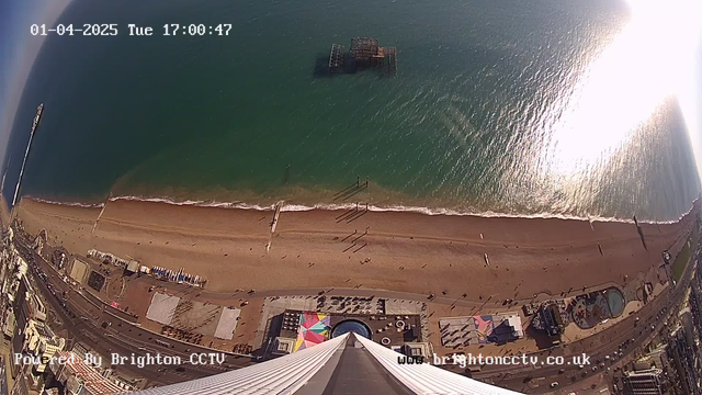 A high-angle view of a beach scene on a clear day. The sandy shore extends horizontally across the bottom of the image, with a few small figures walking along it. Gentle waves lap at the shore, and there is a partially submerged pier in the water further out. In the bottom foreground, various structures, including amusement rides and a pool area, are visible. The sky is mostly clear with hints of clouds, and sunlight reflects off the water, creating a shimmering effect. The date and time are displayed in the top left corner.