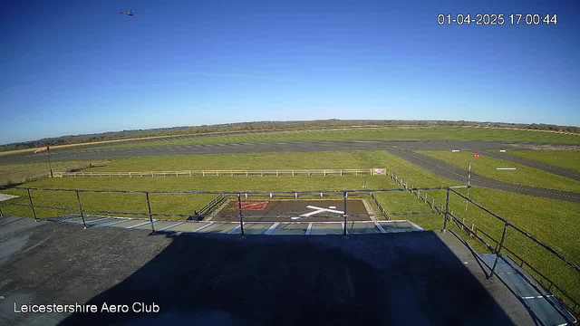 A clear blue sky stretches overhead, with a small helicopter flying in the upper left corner. Below, a wide green field is divided by a white fence, leading to a runway that appears mostly gray. Near the center, a helicopter landing pad marked with a white cross is visible, surrounded by grass and a few signs. The scene is bathed in bright sunlight, with no clouds in the sky.