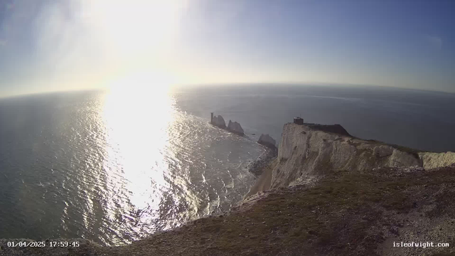 A coastal view featuring cliffs and the ocean. The sun reflects brightly on the water, creating a shimmering effect. In the background, there are several tall rock formations rising from the sea, with one structure resembling a tower on top of a rock. The sky is clear with a few light clouds, and the scene is serene. The foreground shows grassy terrain leading to the edge of the cliffs. The date and time are displayed in the lower left corner.