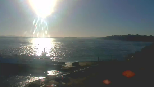 A bright sun shines in a clear sky, reflecting light off the water's surface. In the foreground, a boat is docked near a small pier, while several vehicles are parked on a paved area. The distant shore features a silhouette of hills against the horizon. The scene conveys a tranquil waterfront setting during the day.