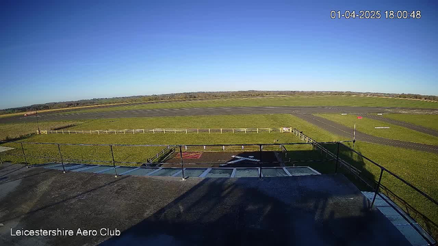 A wide view of an airport runway and surrounding grass fields during daylight. The sky is clear and blue. In the foreground, there is a flat-roofed structure with a railing. A white "X" marking is visible on the ground, possibly indicating a helipad or runway designation. To the left, a red windsock is positioned, indicating wind direction. The area is mostly open with a few scattered runway markings visible on the tarmac.