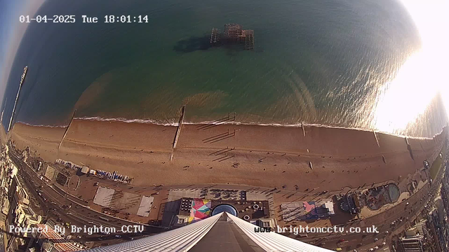 A view from a high vantage point overlooking a sandy beach and the ocean. Below, people are walking along the beach, with shadows cast on the sand. There is a pier extending into the water in the distance, along with remnants of a structure partially submerged. The coastline includes a promenade with colorful buildings and amusement rides in the foreground. The scene is bright, with sunlight reflecting on the water surface.