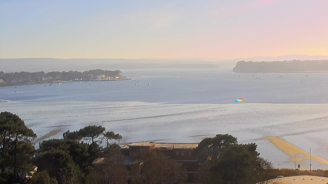A scenic view of a calm body of water during sunset, with gentle waves and a serene sky transitioning from blue to soft hues of pink and orange. In the distance, small boats are scattered on the water, and a coastal landscape with trees and buildings is visible along the shore. The water reflects the colors of the sky, creating a tranquil atmosphere.