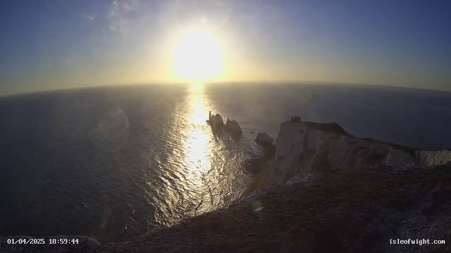 A coastal landscape at sunset, featuring the sun low on the horizon glowing brightly and reflecting off the ocean water. To the right, there are tall white cliffs, and further off in the ocean, a series of rocky islets can be seen. The scene conveys a peaceful setting with a tranquil sea and a clear sky gradually transitioning from blue to warmer tones near the sun.