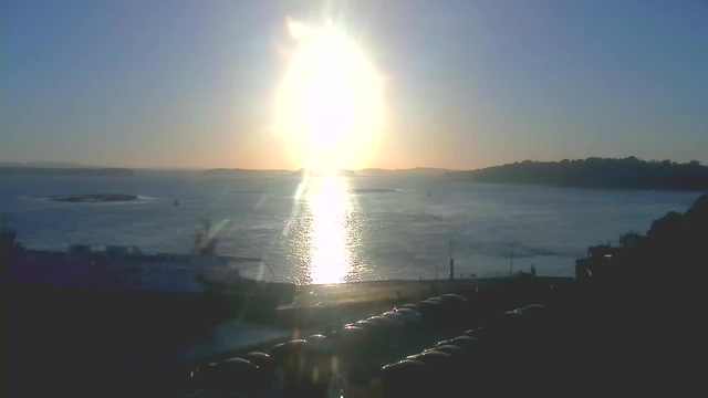 A bright sun rises over a calm body of water, casting a shimmering reflection across the surface. The sky is a gradient of light blue to pale orange near the horizon. In the foreground, a dark silhouette of a ship is partially visible, along with a line of cars parked along the waterfront. The landscape in the background features distant hills and a clear horizon.