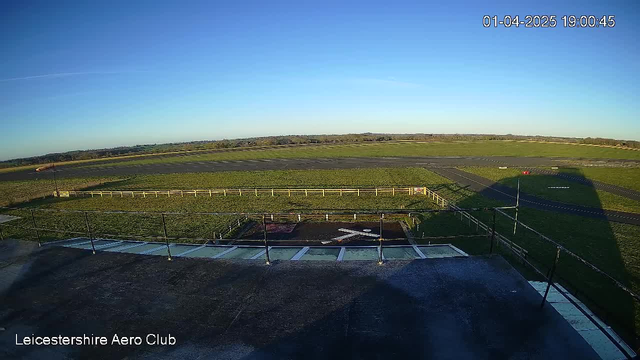 A clear blue sky stretches across the top of the image, with no clouds visible. Below, the scene depicts a wide-open grassy area that encompasses a runway. To the left, there are faint outlines of structures and a few trees in the distance. A white wooden fence lines part of the grassy area, enclosing a smaller dark patch, possibly a runway or landing strip marked with a white cross. The temperature appears cool, and no aircraft are visible on the ground. The time stamp at the top right indicates the image was captured on April 1, 2025, at 19:00:45.