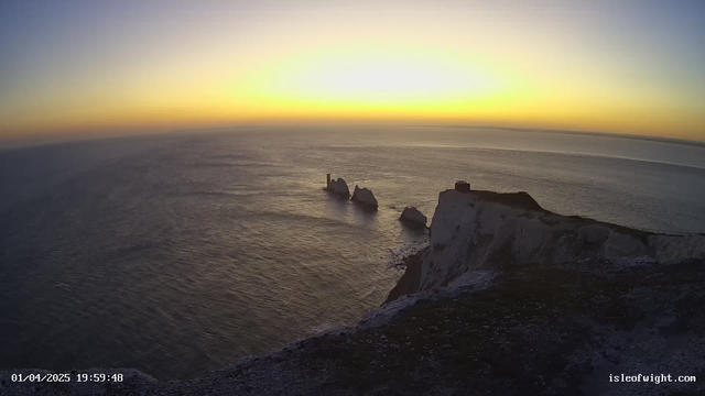 A scenic view of the sea at sunset, with a gradient of colors transitioning from deep blue at the top to warm orange and yellow near the horizon. In the foreground, white cliffs are visible on the right side, with a rocky formation and a small tower or structure on the left. The water reflects the sunset colors, creating a serene atmosphere.