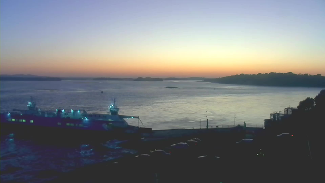 A tranquil waterfront scene during twilight. In the foreground, there is a large boat docked near the shore, illuminated by blue lights. The water reflects the soft colors of the sunset, transitioning from yellow to orange to deep blue in the sky. Silhouettes of hills are visible in the distance, along with a few small boats dotting the water. Cars are parked along the edge of the dock area, contributing to the peaceful atmosphere of the evening.