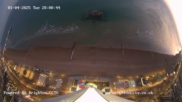 An aerial view of a beach at dusk. The shoreline is visible with gentle waves lapping at the sand. A pier extends into the water, with an old structure visible at its end. The beach is lined with various buildings and features, including colorful lights along the promenade. The scene is illuminated by artificial lighting, creating a warm glow. The date and time are displayed at the top of the image.