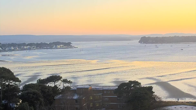 A serene view of a waterbody at sunset, with soft, warm tones in the sky ranging from light orange to pastel blue. Silhouetted trees and buildings are in the foreground, while the water reflects the colors of the sky. Several small boats can be seen moving gently on the water, and distant hills are visible along the horizon. The scene conveys a calm and peaceful atmosphere.