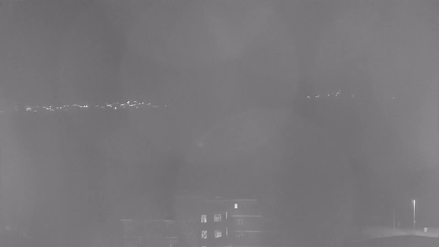 A dimly lit scene captured by a webcam showing a hazy view of lights in the distance against a dark background. A building with windows is partially visible at the bottom of the image, and there are faint points of light along the horizon, indicating distant structures or lights. The overall atmosphere is misty and lacks clarity.