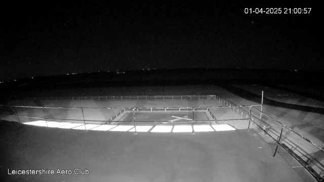 A dark, nighttime view from a webcam shows a wide, flat outdoor area with a rectangular shape towards the center, which is slightly illuminated. The scene is mostly shadowed, with some distant lights visible in the background. A metal railing surrounds the illuminated area, suggesting a boundary. The timestamp indicates the image was captured on April 1, 2025, at 9:00 PM. The bottom left corner includes the text "Leicestershire Aero Club".