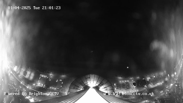 A black and white image from a webcam showing a nighttime scene. The view appears to be a wide angle from a height, with faint lights visible in the distance. The bottom of the image features a bright white area that draws the eye, while the upper part is darker with some indistinct shapes. The time stamp shows the date as January 4, 2025, at 21:01:23. The image is labeled “Powered By Brighton CCTV” in the bottom left corner.