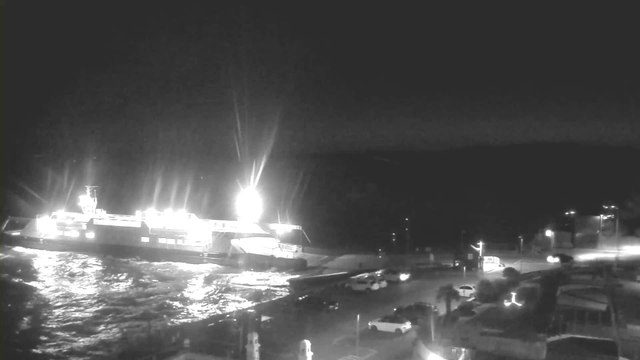 A bright white ferry docked at night, illuminated by strong lights. Waves gently ripple in the water around the ferry. In the foreground, there are several parked cars and streetlights casting a soft glow. The background features dark hills, blending into the night sky.