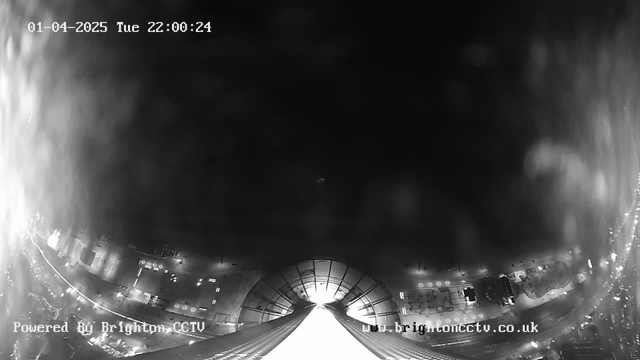 A black and white image from a webcam shows a nighttime cityscape. In the foreground, there is a large structure with a curved design, possibly a dome or roof, leading toward a bright light source in the distance, suggesting a far-off illumination. The bottom of the image captures parts of a road or pathway, with faint outlines of vehicles or objects that indicate urban activity. The scene is mostly dark, with scattered light spots along the edges, hinting at streetlights or buildings in the surroundings.