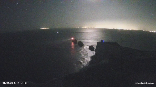 A dimly lit coastal scene at night, featuring a rocky shoreline and the sea. In the foreground, several dark rocks emerge from the water. To the left, a small boat with a red light is visible, while some lights from a distant shoreline can be seen in the background. The sky is dark with a few scattered stars. A blue light is visible on the rocky cliff at the right side of the image. The atmosphere is tranquil and somewhat mysterious.
