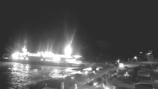 A brightly lit ferry docked at a harbor during the night. The water reflects the lights from the ferry and surroundings. Various vehicles are parked along a street next to the harbor, and a few more cars are moving on the road to the right. The scene is illuminated by both the ferry's lights and street lamps.