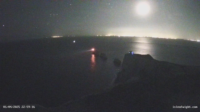 A night scene over the ocean, featuring a rocky coastline with cliffs on the right side. The sky is dark with stars visible and a bright full moon shining above. On the water, there are two boats: one is emitting a red light, and the other is farther in the distance, showing a white light. In the background, a faint glow from a distant shoreline can be seen. The date and time are shown in the bottom left corner.