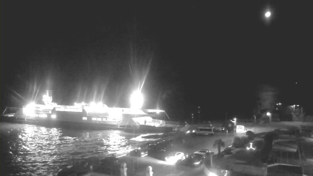 A brightly lit ferry docked at a harbor at night, with lights reflecting on dark water. Several parked vehicles are visible near the dock, and a bright moon is shining in the sky. The scene has a somber yet illuminated atmosphere, with shadows cast by the lights.